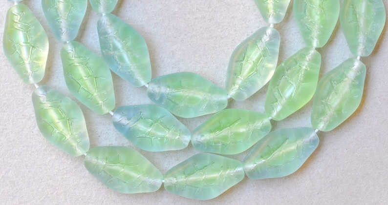 May include: Two strands of translucent leaf-shaped beads in shades of green and blue. The beads are strung on a white cord, and each bead has a detailed leaf vein pattern. The beads are approximately 1 inch long.