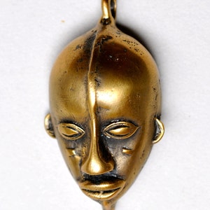 May include: A gold-colored metal pendant in the shape of a stylized human face. The face has closed eyes, a prominent nose, and a slightly open mouth.