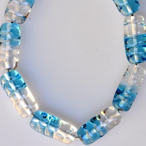 Puede incluir: Un collar hecho de cuentas de vidrio azul y transparente. Las cuentas son largas y rectangulares, con un patrón ondulado. El collar está enhebrado en una cadena plateada.