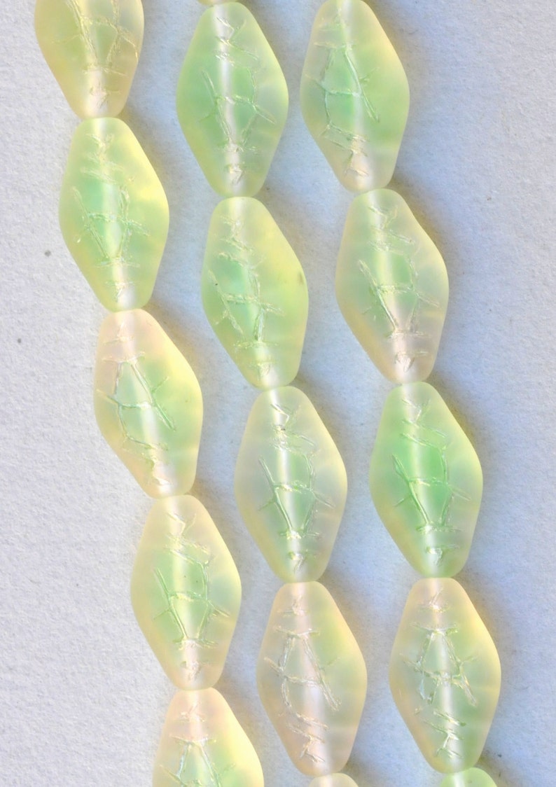 May include: Close-up of several strands of light green, diamond-shaped beads. Each bead has a frosted appearance with a subtle, internal pattern. The beads are strung together, creating a delicate and elegant look.