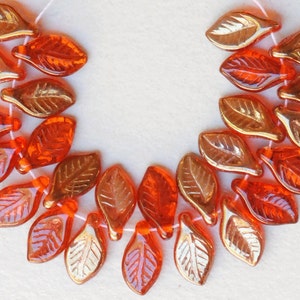May include: A strand of orange and gold glass leaf beads. The beads are strung on a white thread and are arranged in a semi-circle.