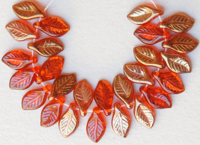 May include: A strand of orange and gold glass leaf beads. The beads are strung on a white thread and are arranged in a semi-circle.