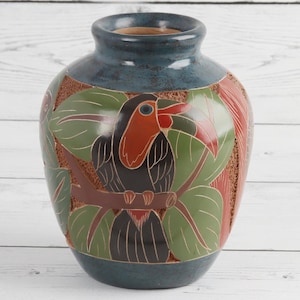 May include: A ceramic vase featuring a hand-painted toucan perched on a branch amidst green leaves. The vase has a dark blue rim and base, with a textured, reddish-brown background behind the bird. The toucan has a black body and a large orange beak.