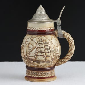May include: A ceramic beer stein featuring a relief of a sailing ship at sea with buildings in the background. The stein has a metallic lid with a thumb lever and a handle shaped like a rope. The base includes the text "Bark Staysail Schooner".