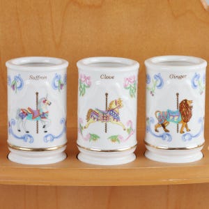 May include: Three white ceramic cylindrical containers featuring carousel animal illustrations. The first shows a unicorn labeled Saffron, the second a horse labeled Clove, and the third a lion labeled Ginger. Each has gold trim and floral accents.