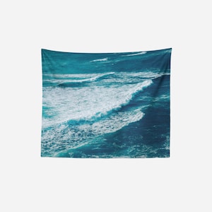 May include: A wall tapestry featuring an aerial view of ocean waves. The waves are a mix of deep and light blues, with white foamy crests. The tapestry is rectangular and hangs against a white background.