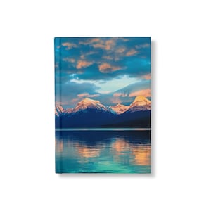 May include: A scenic landscape photograph featuring a mountain range with snow-capped peaks reflected in a still lake at sunset. The sky is a vibrant blue with pink and orange clouds.