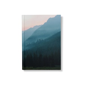 May include: A misty mountain landscape with a forest of evergreen trees in the foreground. The sky is a soft pink and blue, and the mountains are a deep blue.