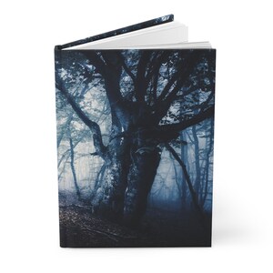 May include: Hardcover journal featuring a dark, atmospheric forest scene. The cover displays a large, gnarled tree in a misty, blue-toned forest. The open journal reveals white pages, suggesting a space for writing or sketching.