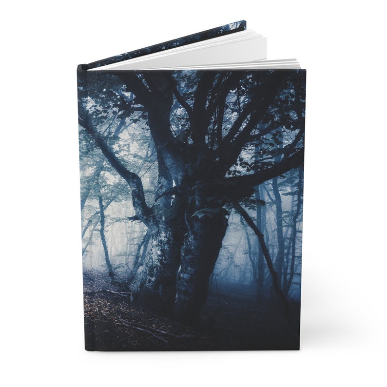 May include: Hardcover journal featuring a dark, atmospheric forest scene. The cover displays a large, gnarled tree in a misty, blue-toned forest. The open journal reveals white pages, suggesting a space for writing or sketching.