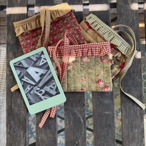 May include: A light green e-reader displaying a screen of scattered letters alongside three quilted fabric pouches. The pouches feature floral patterns in red, green, and tan, each with a ruffled top edge and matching fabric ties for closure.