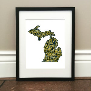 May include: A framed print of a map of Michigan, with the state outline filled in with yellow and the cities and towns within the state outlined in blue. The cities and towns are spelled out in a stylized font.