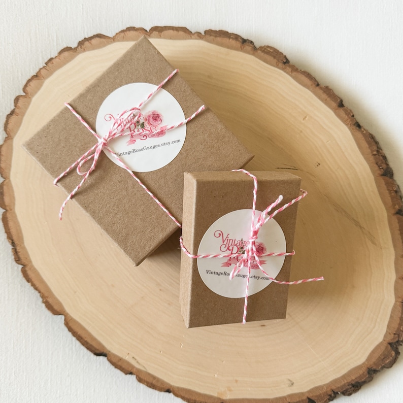 May include: Two small brown gift boxes tied with red and white twine. Each box has a white circle sticker with a pink floral design and the text "VintageRoseGauges.etsy.com".