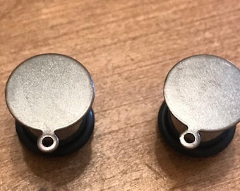 Puede incluir: Un par de dilatadores de oreja de metal plateado con un diseño circular y un pequeño bucle en la parte inferior.