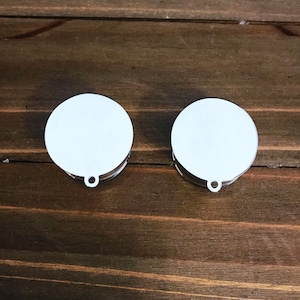 May include: Two white round blank baubles with a silver ring at the top. The baubles are on a brown wooden surface.