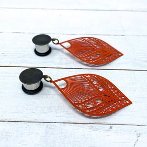 May include: A pair of orange metal earrings with a delicate, intricate design. The earrings are shaped like a leaf and have a dangling charm. The earrings are attached to a silver metal plug.