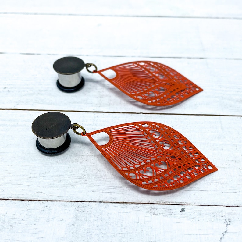 May include: A pair of orange metal earrings with a delicate, intricate design. The earrings are shaped like a leaf and have a dangling charm. The earrings are attached to a silver metal plug.