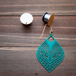 May include: A pair of teal blue earrings with a delicate, intricate design. The earrings are made of metal and have a gold-tone finish. The earrings are hanging from a gold-tone hook.
