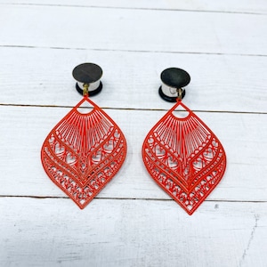 May include: A pair of red metal earrings with a delicate, intricate design. The earrings are shaped like a teardrop and feature a filigree pattern with hearts. The earrings are attached to a silver-colored post.