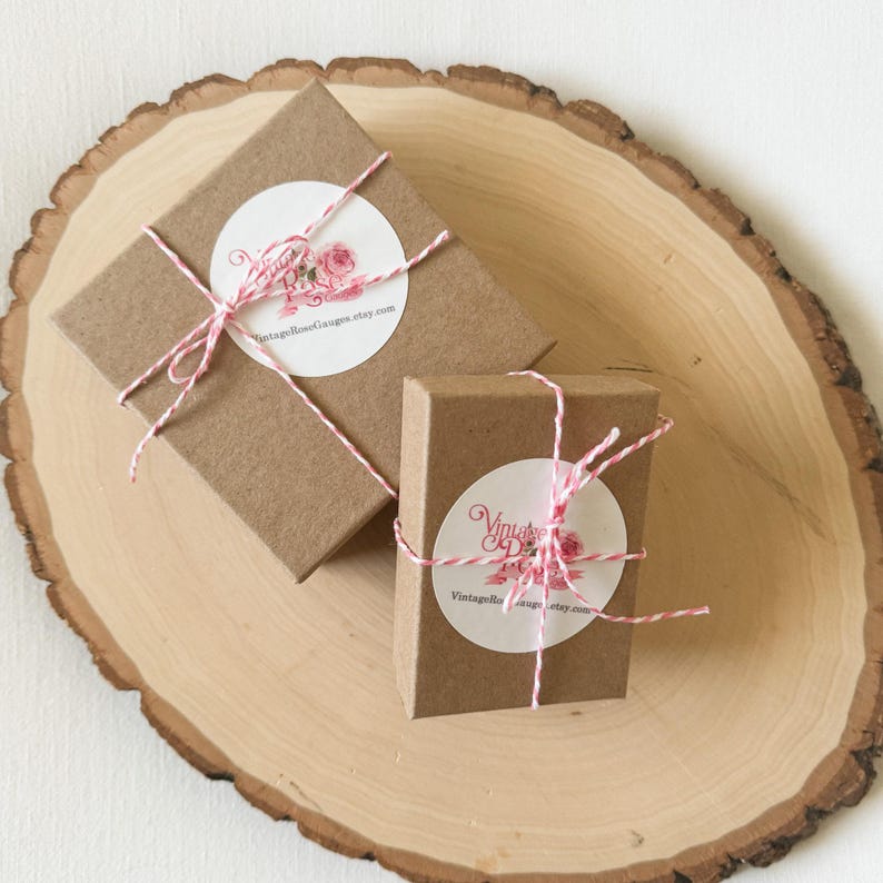 May include: Two small brown boxes with white labels and red and white twine. The labels have a pink rose design and the text "VintageRoseDesigns.etsy.com".