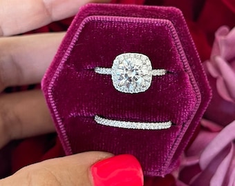 May include: A close-up of a silver ring with a large, square-shaped diamond in a halo setting. The ring is in a red velvet ring box.