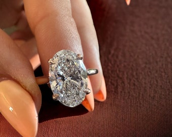 May include: A gold ring featuring a large, oval-cut diamond. The diamond is clear and sparkling, set with four prongs. The ring is held up against a brown background, with a hand and orange nail polish visible.