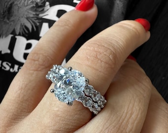 May include: A silver ring set with a large, clear, oval-shaped diamond. The ring has a double band with smaller diamonds set around the main stone.