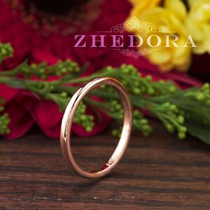May include: A thin rose gold band ring, lying on a wooden surface with a blurred background of red and yellow flowers.