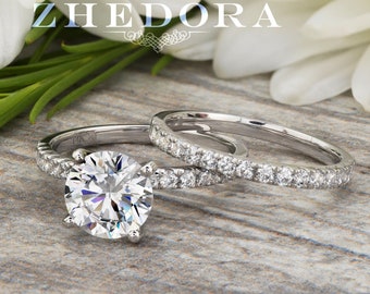 May include: A set of two silver wedding bands with a large round diamond in the centre of the engagement ring. The rings are on a wooden surface with white flowers in the background.