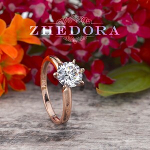 May include: A rose gold engagement ring with a round, clear diamond center stone. The ring is surrounded by colorful flowers.