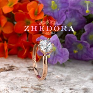 May include: A rose gold engagement ring with a round, clear center stone and a six-prong setting. The ring is surrounded by colorful flowers.