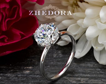 May include: A silver solitaire engagement ring with a large, round, clear diamond. The ring is set against a background of pink and white flowers.