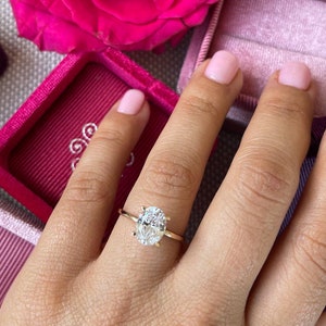 May include: A close-up of a hand wearing a gold ring with an oval-shaped diamond. The ring is set in a simple band. The hand is resting on a pink velvet box.