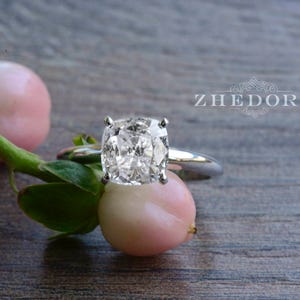 May include: A silver ring with a large, clear, oval-shaped diamond. The ring is set on a wooden surface with pink flowers.