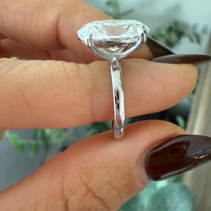 May include: A close-up of a silver ring with a large, clear, cushion-cut gemstone. The ring is held up, showcasing the intricate setting and the brilliance of the stone. The band is thin and polished, reflecting light.