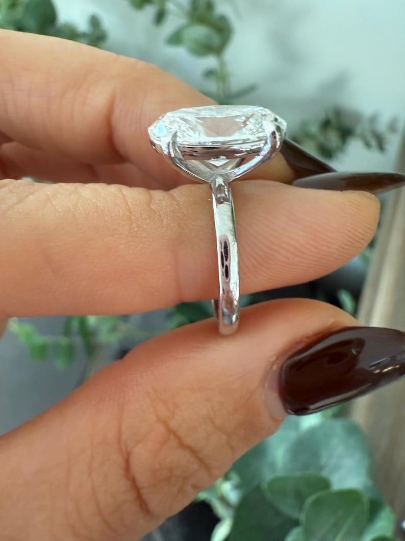 May include: A close-up of a silver ring with a large, clear, cushion-cut gemstone. The ring is held up, showcasing the intricate setting and the brilliance of the stone. The band is thin and polished, reflecting light.
