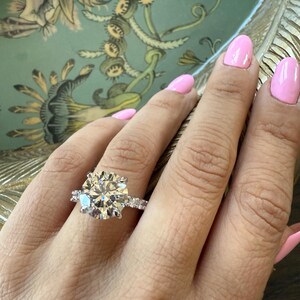 K&ouml;nnte beinhalten: Ein Diamant-Verlobungsring mit einem gro&szlig;en, rund geschliffenen Mittelstein, umgeben von kleineren Diamanten. Der Ring, gefasst in einem silbernen Band, wird auf einer Hand mit rosa Nagellack gezeigt. Der Hintergrund hat ein Blumenmuster.