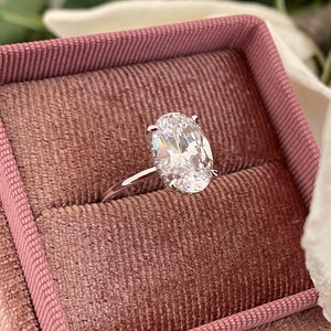 Puede incluir: Un anillo de plata con un gran diamante de forma ovalada en una caja de anillo de terciopelo rosa.