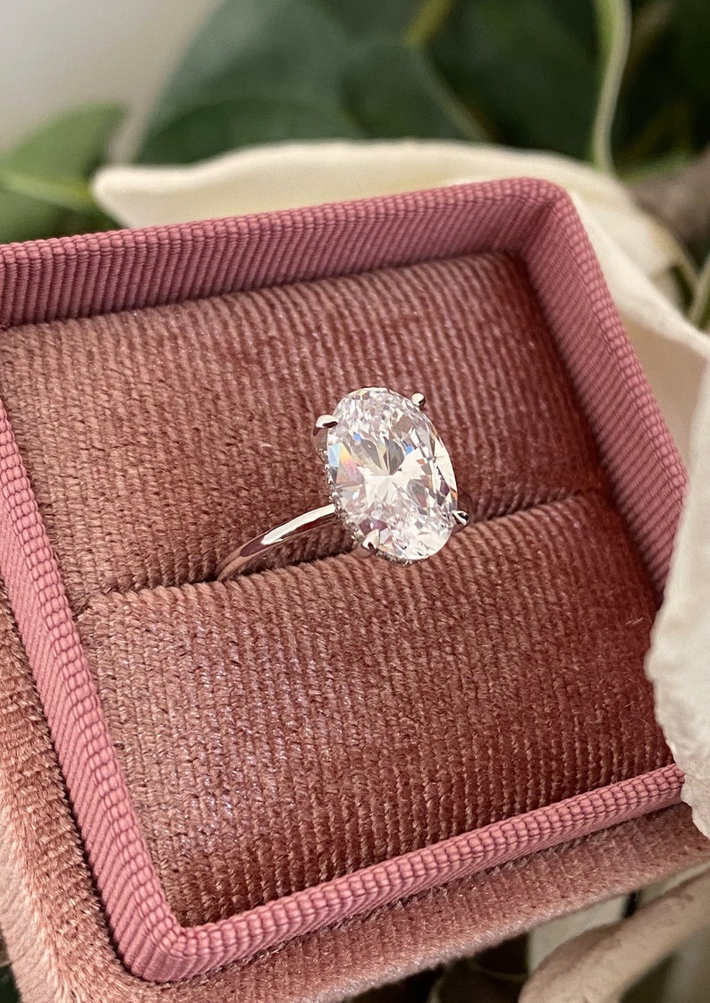 Puede incluir: Un anillo de plata con un gran diamante de forma ovalada en una caja de anillo de terciopelo rosa.