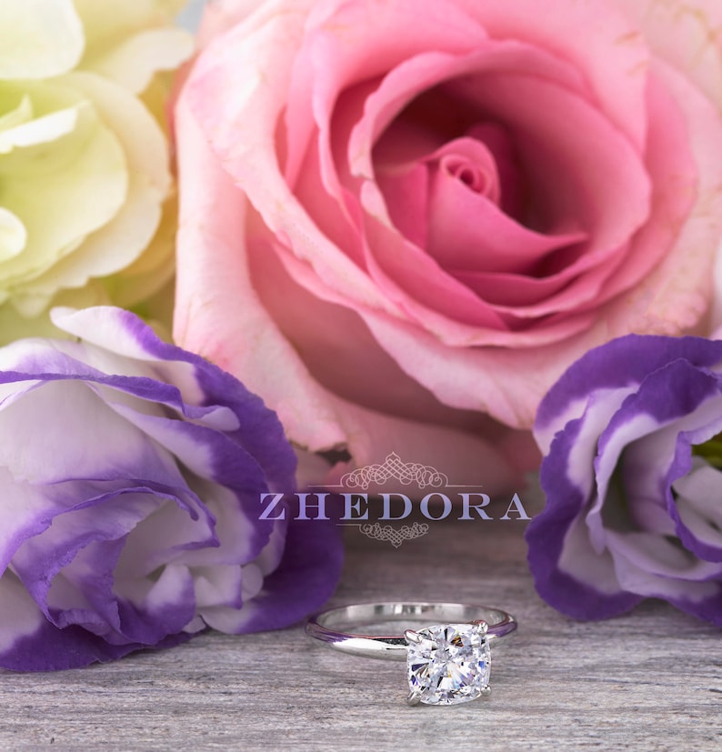 May include: A silver engagement ring with a round, clear diamond set in a simple band. The ring is surrounded by pink and purple flowers.
