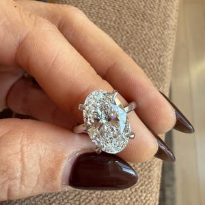 May include: A silver ring featuring a large, oval-cut diamond. The diamond is clear and sparkling, set with four prongs. The ring is held up against a neutral background.