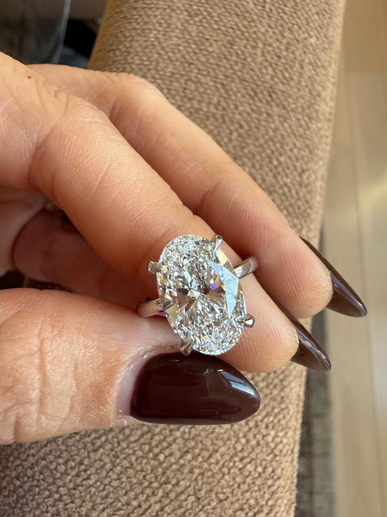 May include: A silver ring featuring a large, oval-cut diamond. The diamond is clear and sparkling, set with four prongs. The ring is held up against a neutral background.