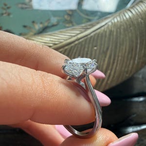 Pu&ograve; includere: Un anello d'argento con un grande diamante rotondo e trasparente come elemento centrale. Il diamante &egrave; tenuto in posizione da pi&ugrave; griffe. La fascia dell'anello &egrave; liscia e lucida, riflettendo la luce. L'anello &egrave; tenuto dalle dita di una persona.