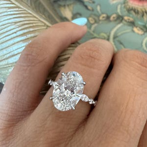 May include: A sparkling diamond ring with an oval-cut center stone and smaller accent stones. The ring is set in a silver band and is displayed on a hand with a light blue manicure. The ring is a piece of fine jewelry.