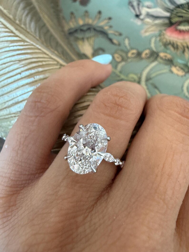 May include: A sparkling diamond ring with an oval-cut center stone and smaller accent stones. The ring is set in a silver band and is displayed on a hand with a light blue manicure. The ring is a piece of fine jewelry.