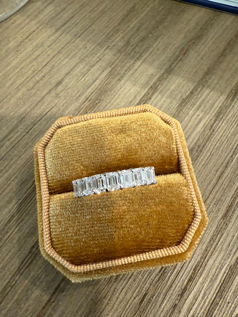 May include: A diamond eternity band ring with emerald cut stones set in a white gold band. The ring is displayed in a velvet ring box.