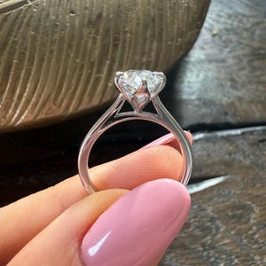 Pu&ograve; includere: Primo piano di un anello di fidanzamento con diamanti. L'anello presenta un grande diamante taglio rotondo incastonato in una fascia in oro bianco o platino. La fascia ha un delicato design a gambo diviso. L'anello &egrave; tenuto su uno sfondo sfocato.