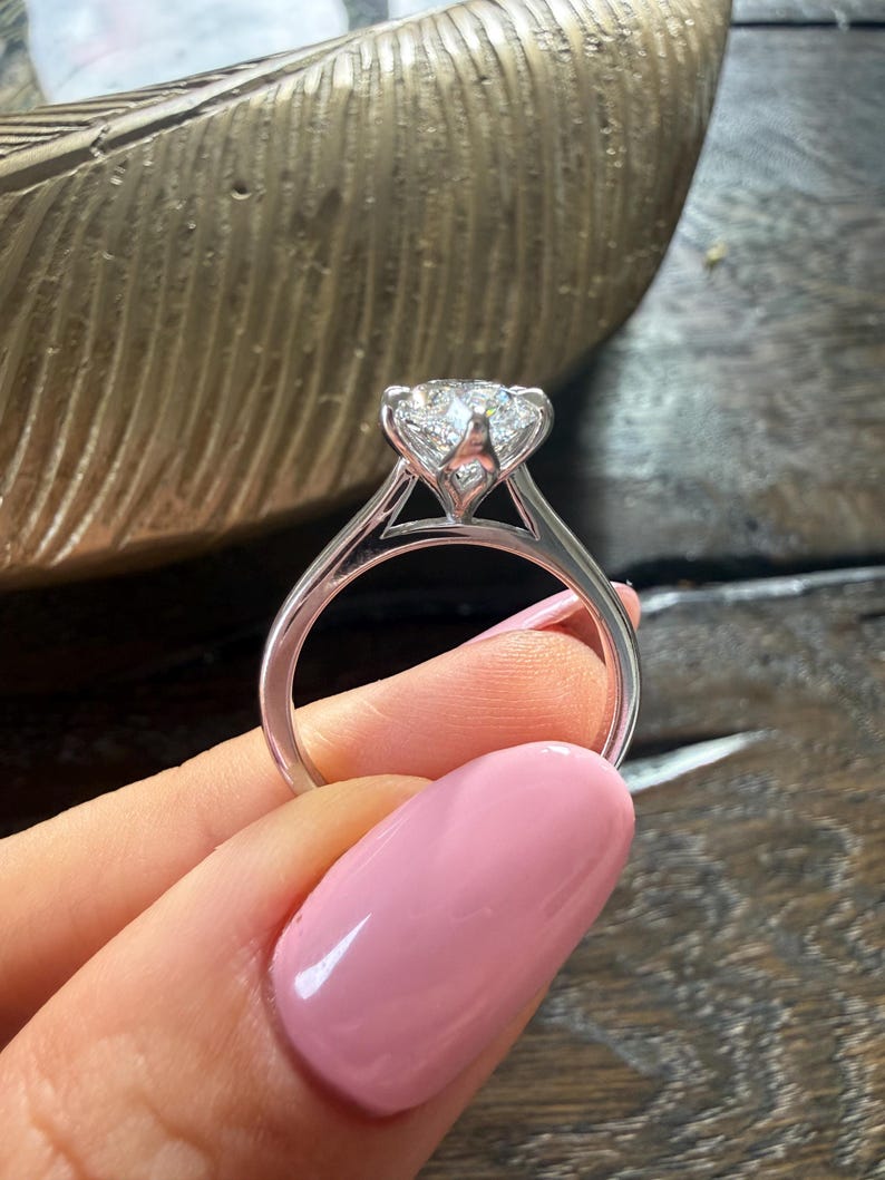 Pu&ograve; includere: Primo piano di un anello di fidanzamento con diamanti. L'anello presenta un grande diamante taglio rotondo incastonato in una fascia in oro bianco o platino. La fascia ha un delicato design a gambo diviso. L'anello &egrave; tenuto su uno sfondo sfocato.