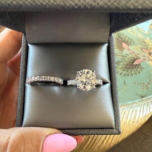 K&ouml;nnte beinhalten: Ein zweiteiliges Ring-Set in einer grauen Ringschatulle. Der Verlobungsring hat einen gro&szlig;en, rund geschliffenen Diamanten mit kleineren Akzentsteinen. Der Ehering ist ein zartes Eternity-Band mit kleinen, funkelnden Steinen. Die Schatulle wird von einer Hand mit rosa Nagellack gehalten.