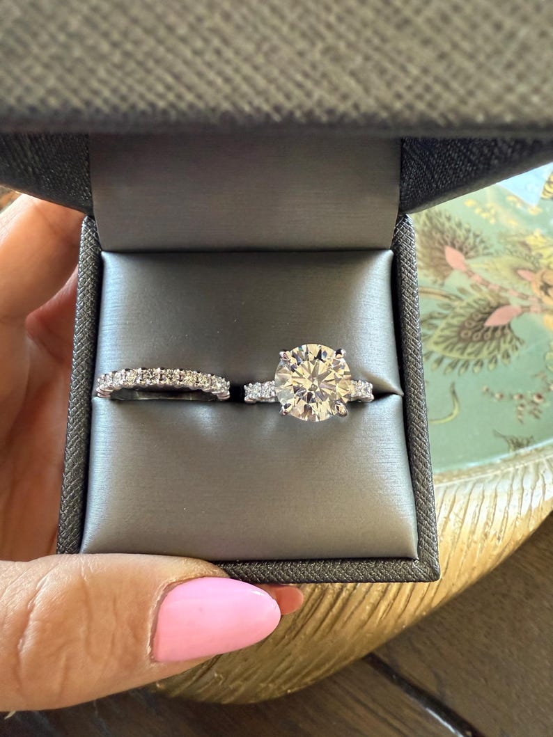 K&ouml;nnte beinhalten: Ein zweiteiliges Ring-Set in einer grauen Ringschatulle. Der Verlobungsring hat einen gro&szlig;en, rund geschliffenen Diamanten mit kleineren Akzentsteinen. Der Ehering ist ein zartes Eternity-Band mit kleinen, funkelnden Steinen. Die Schatulle wird von einer Hand mit rosa Nagellack gehalten.